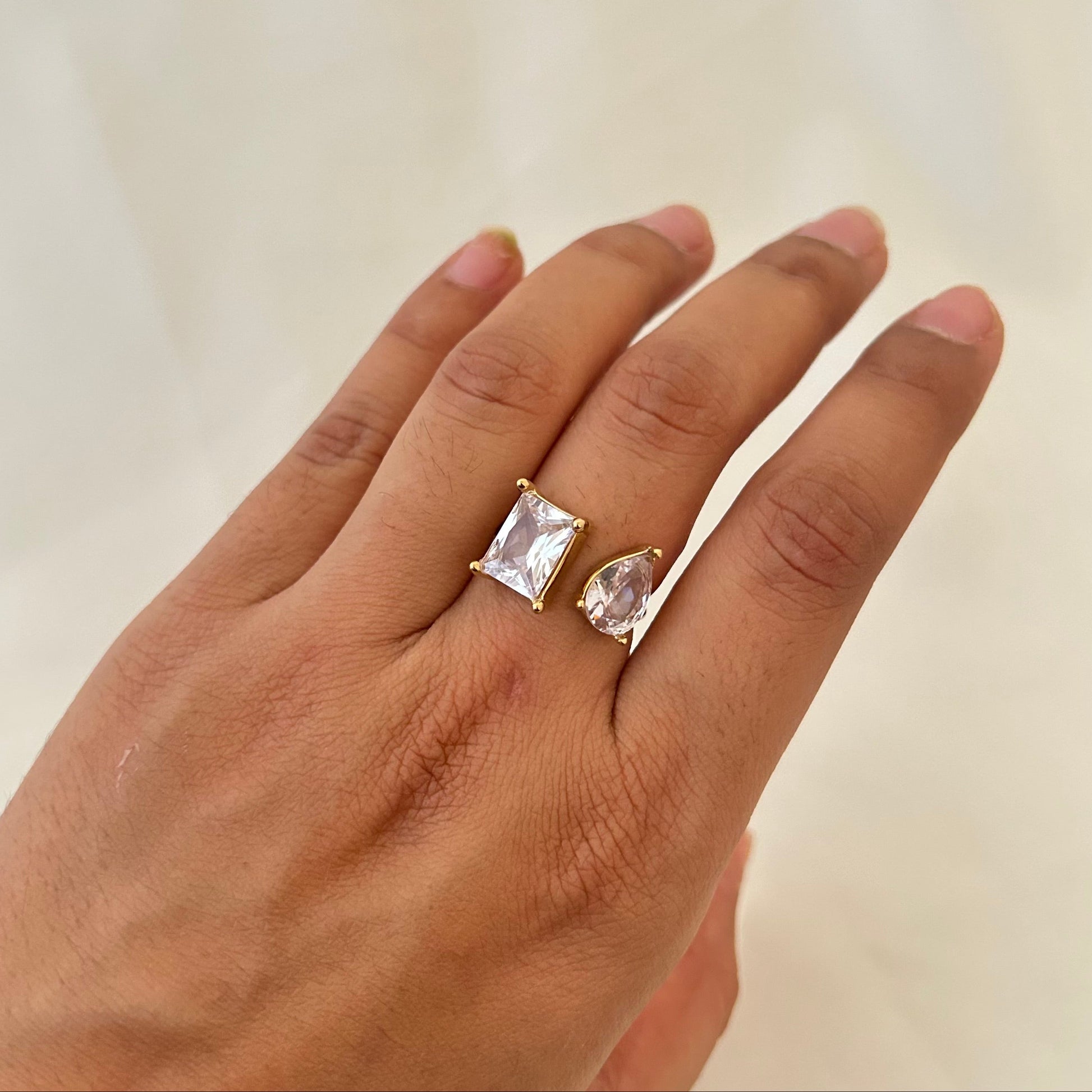 Hand wearing a gold open ring with two clear gemstones, one rectangular and one teardrop-shaped.