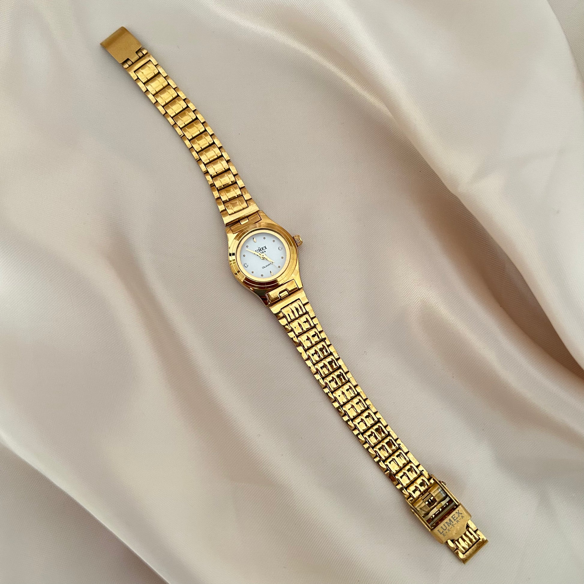 Round gold wristwatch with minimal dial and metallic strap on satin fabric background.