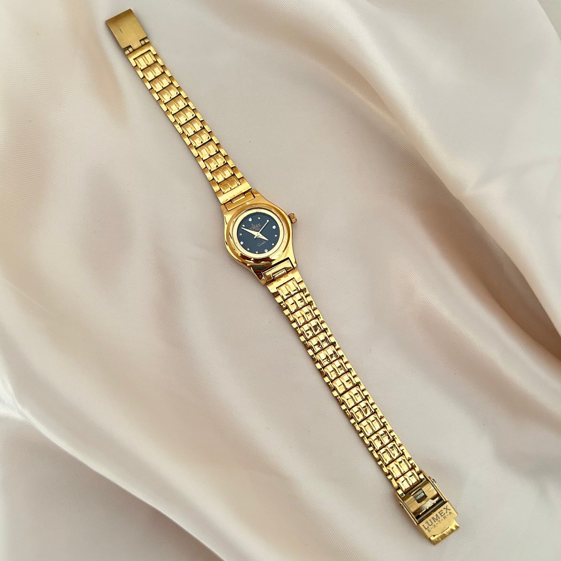 Round gold wristwatch with minimal dial and metallic strap on satin fabric background.