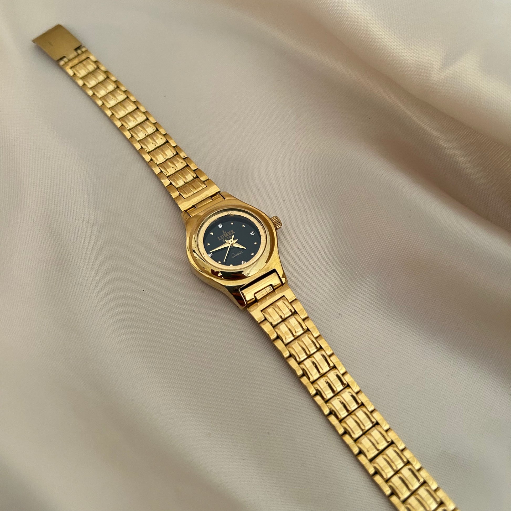 Round gold wristwatch with minimal dial and metallic strap on satin fabric background.