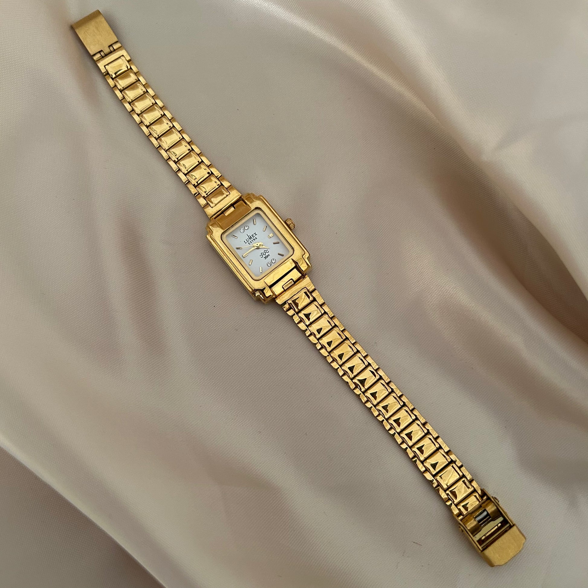 Square gold dial wristwatch with bold strap design on neutral satin background.