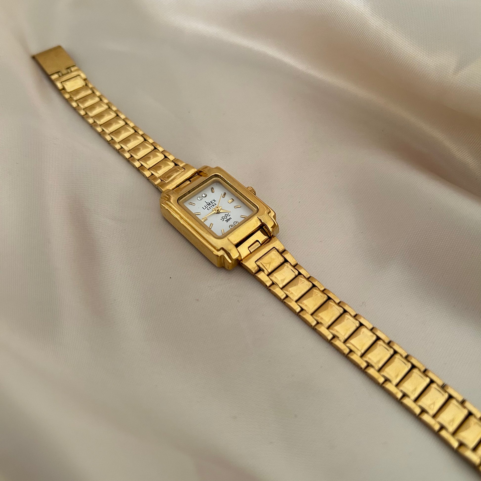 Square gold dial wristwatch with bold strap design on neutral satin background.