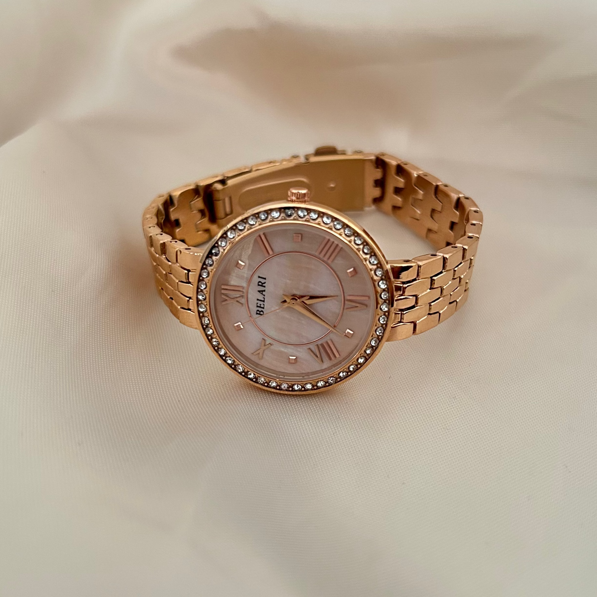 Rose gold wristwatch with Roman numeral dial and crystal studded bezel on soft satin background.
