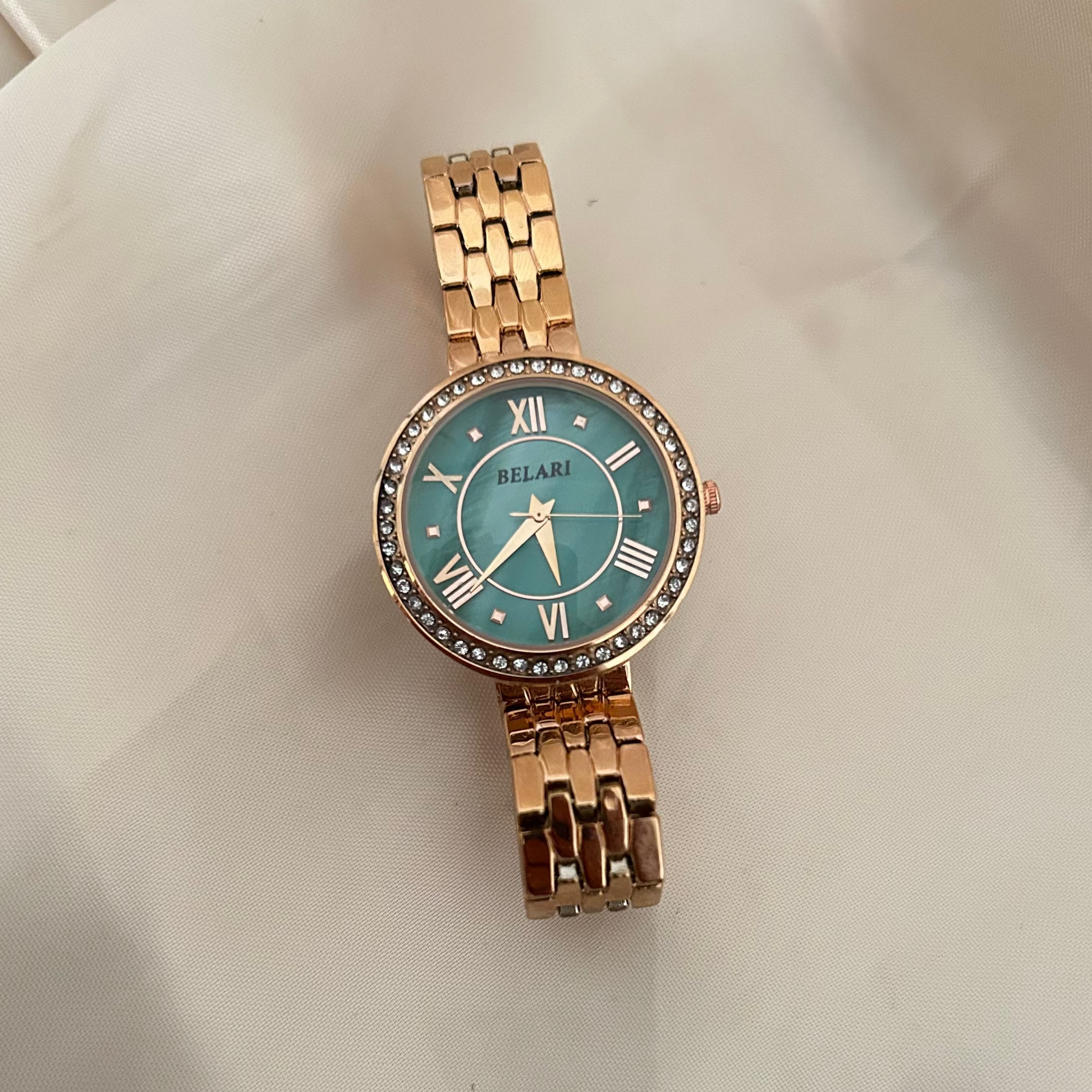 Sea green wristwatch with Roman numeral dial and crystal studded bezel on soft satin background.