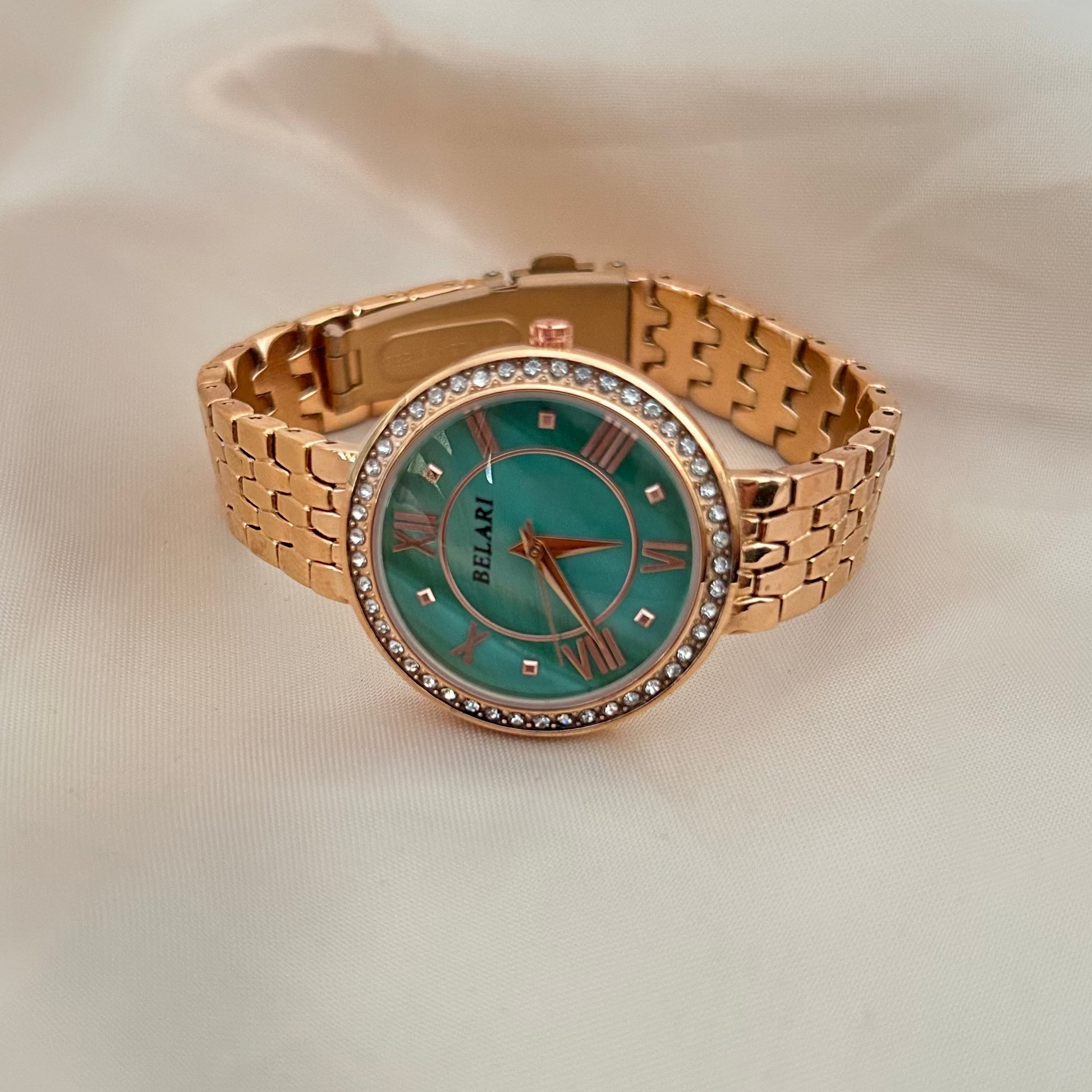 Sea green wristwatch with Roman numeral dial and crystal studded bezel on soft satin background.