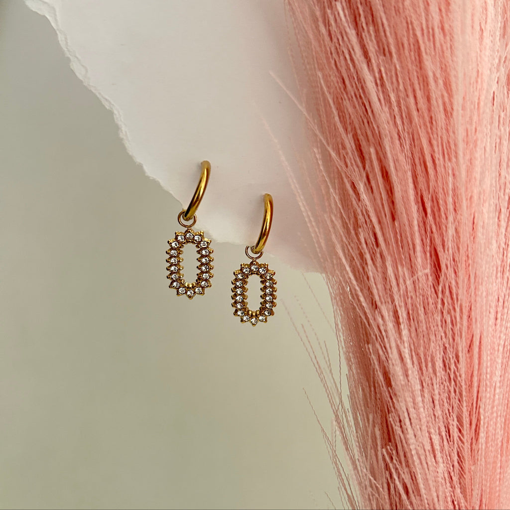 Gold oval drop earrings with small crystal stones arranged in a halo pattern, hanging from delicate gold hoops on a soft neutral background.