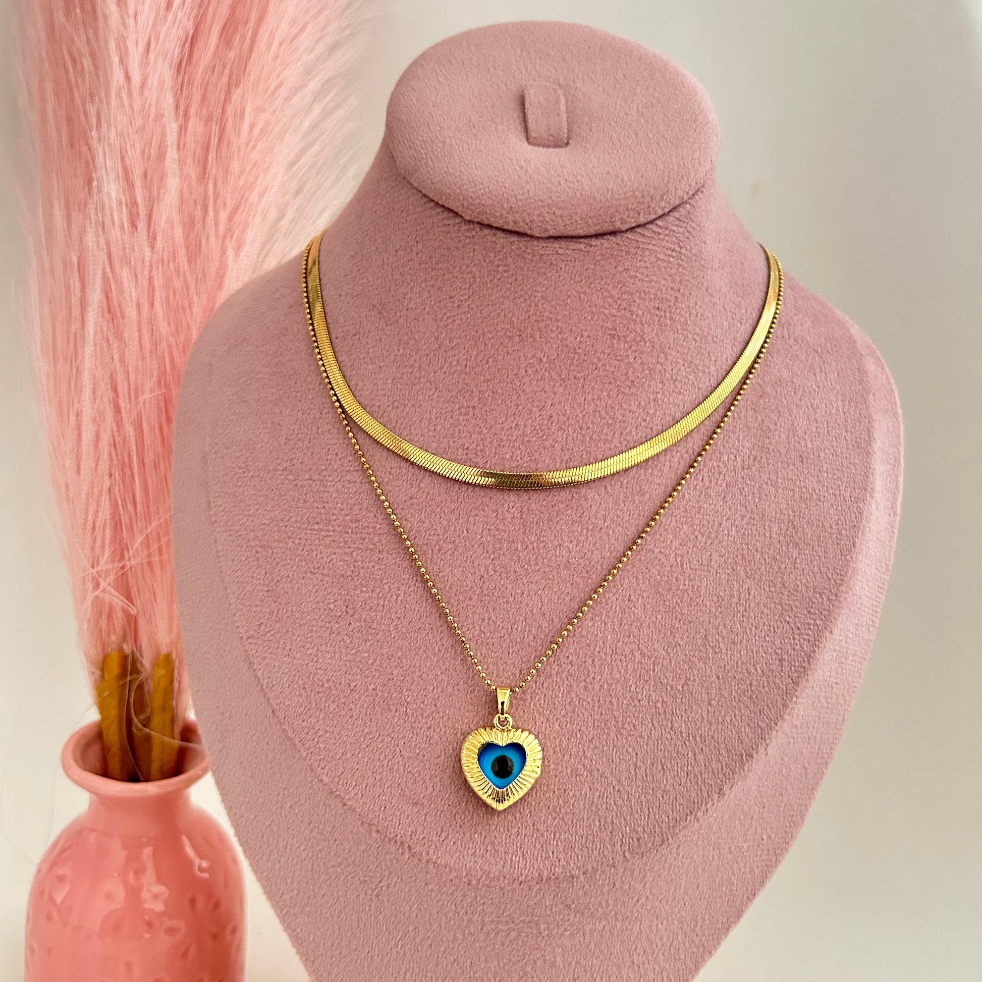 Layered gold necklace with blue evil eye heart pendant on soft pink background