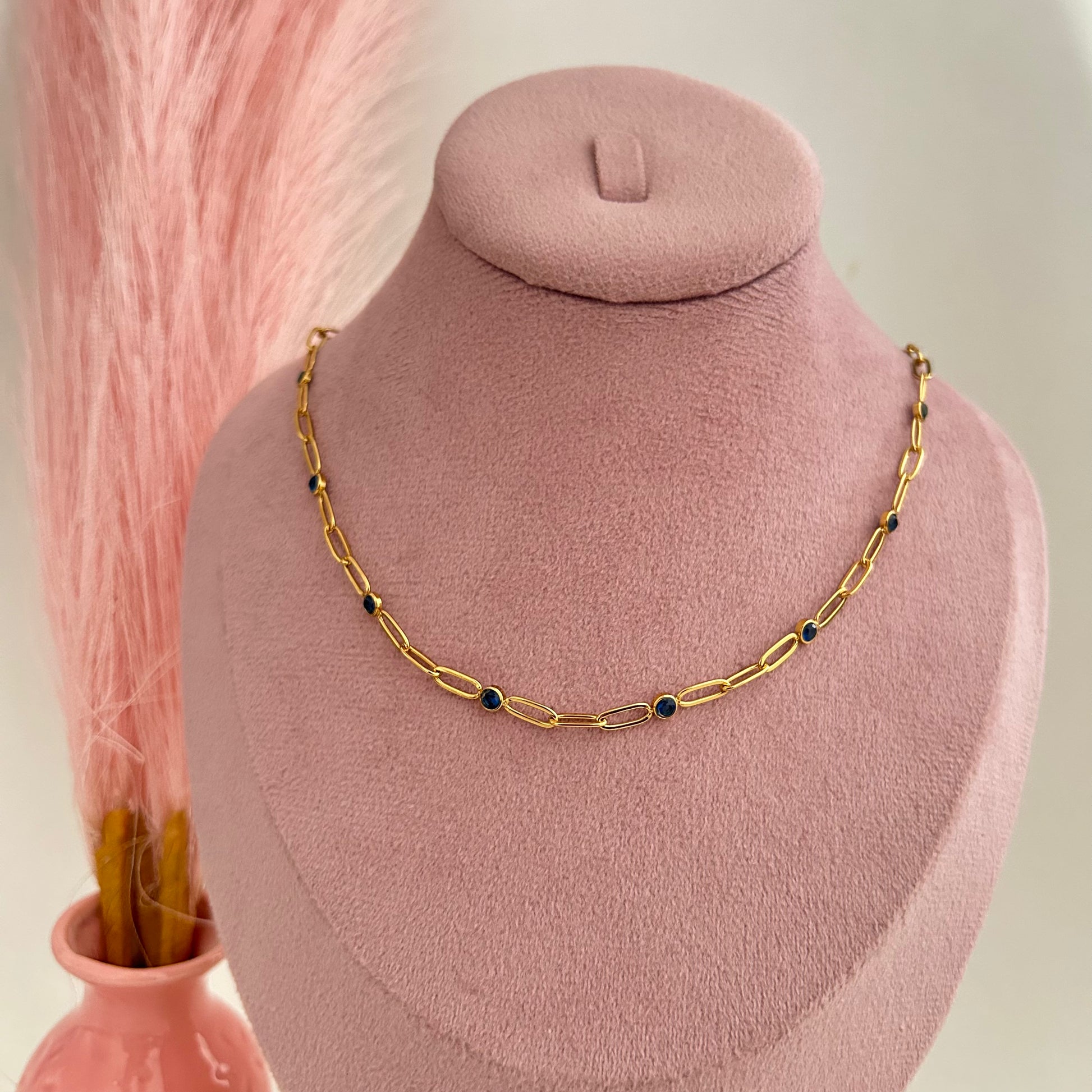 Gold link chain necklace with small blue evil eye charms on a pink display stand