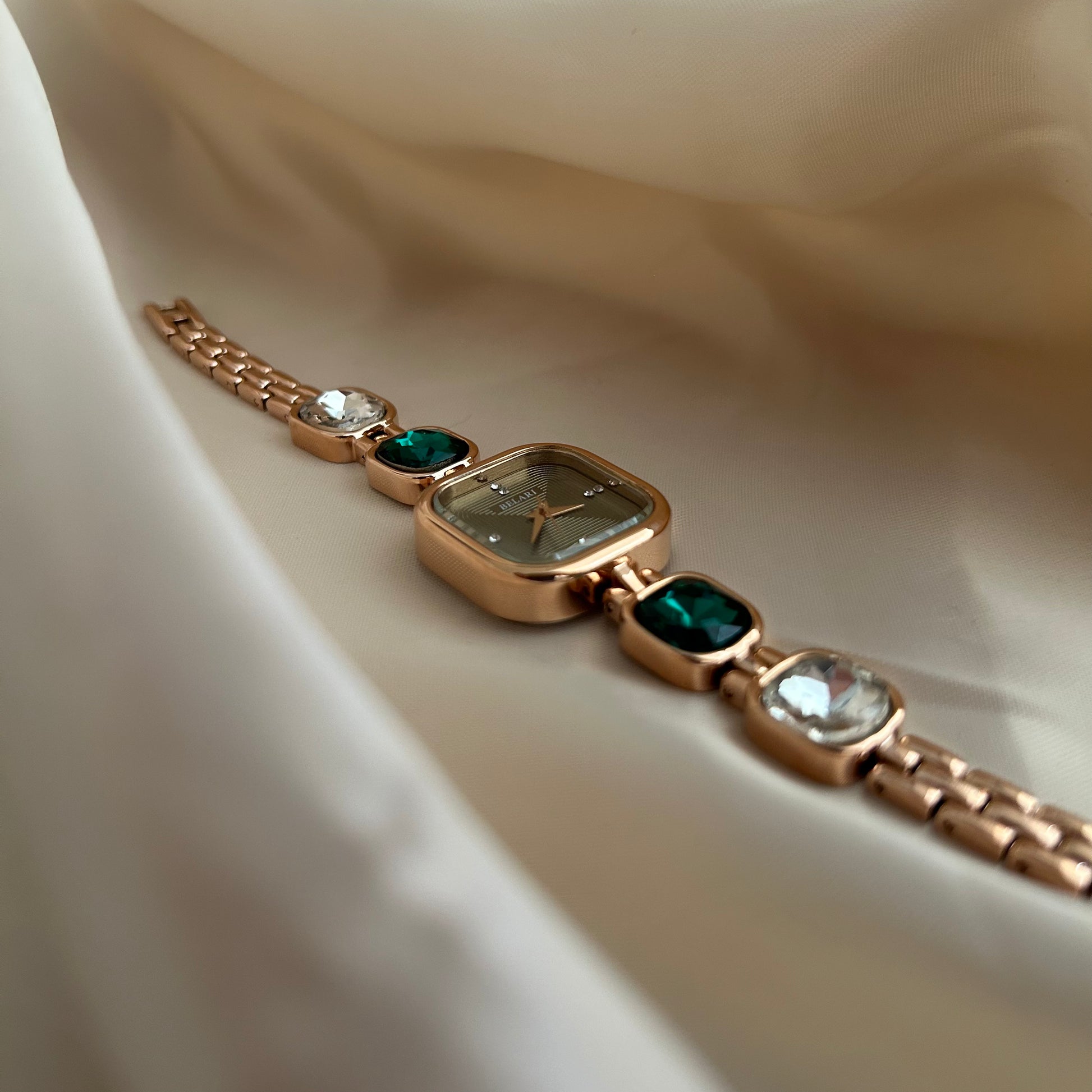 Gold wristwatch with gemstone bracelet links and square dial on satin background.