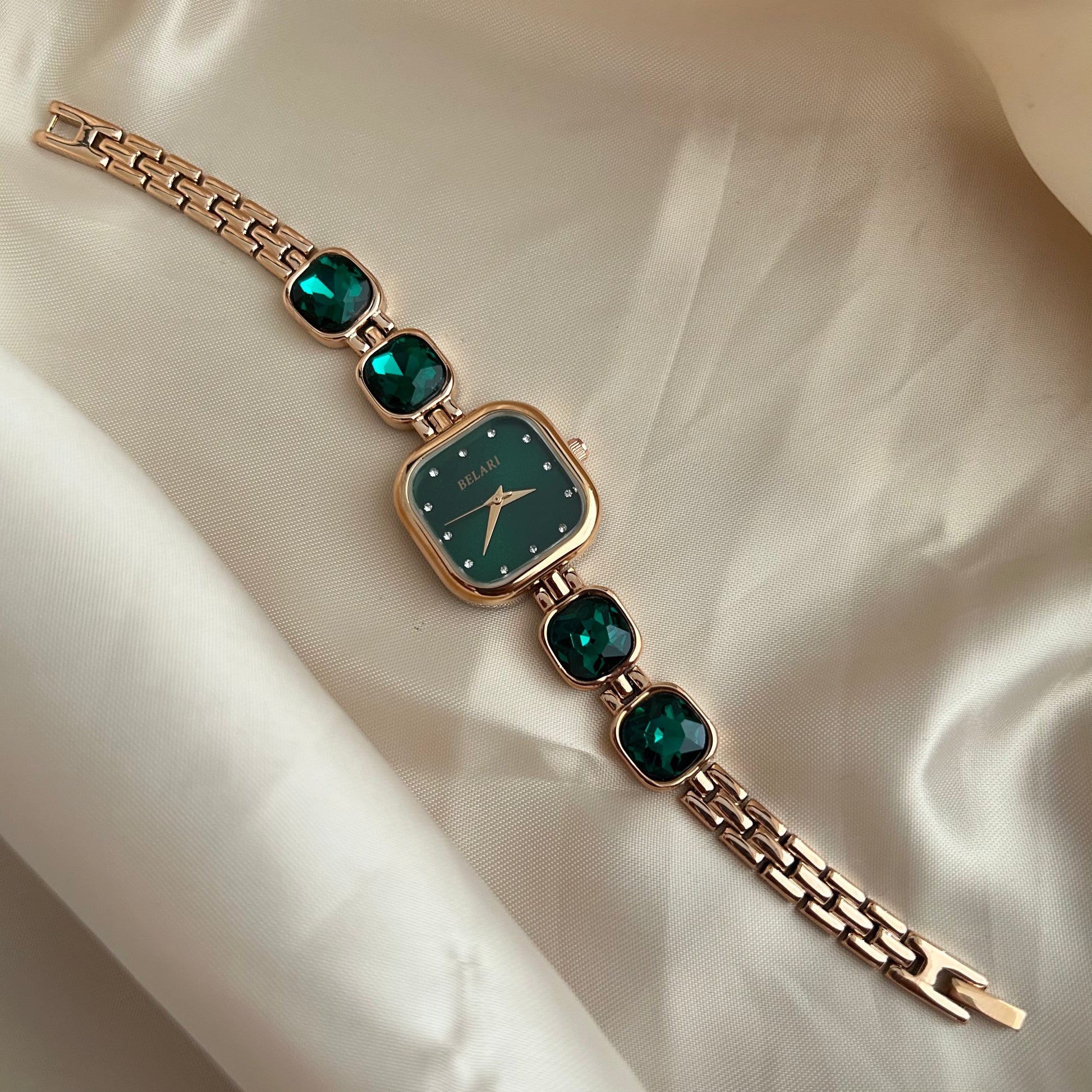 Gold wristwatch with gemstone bracelet links and square dial on satin background.