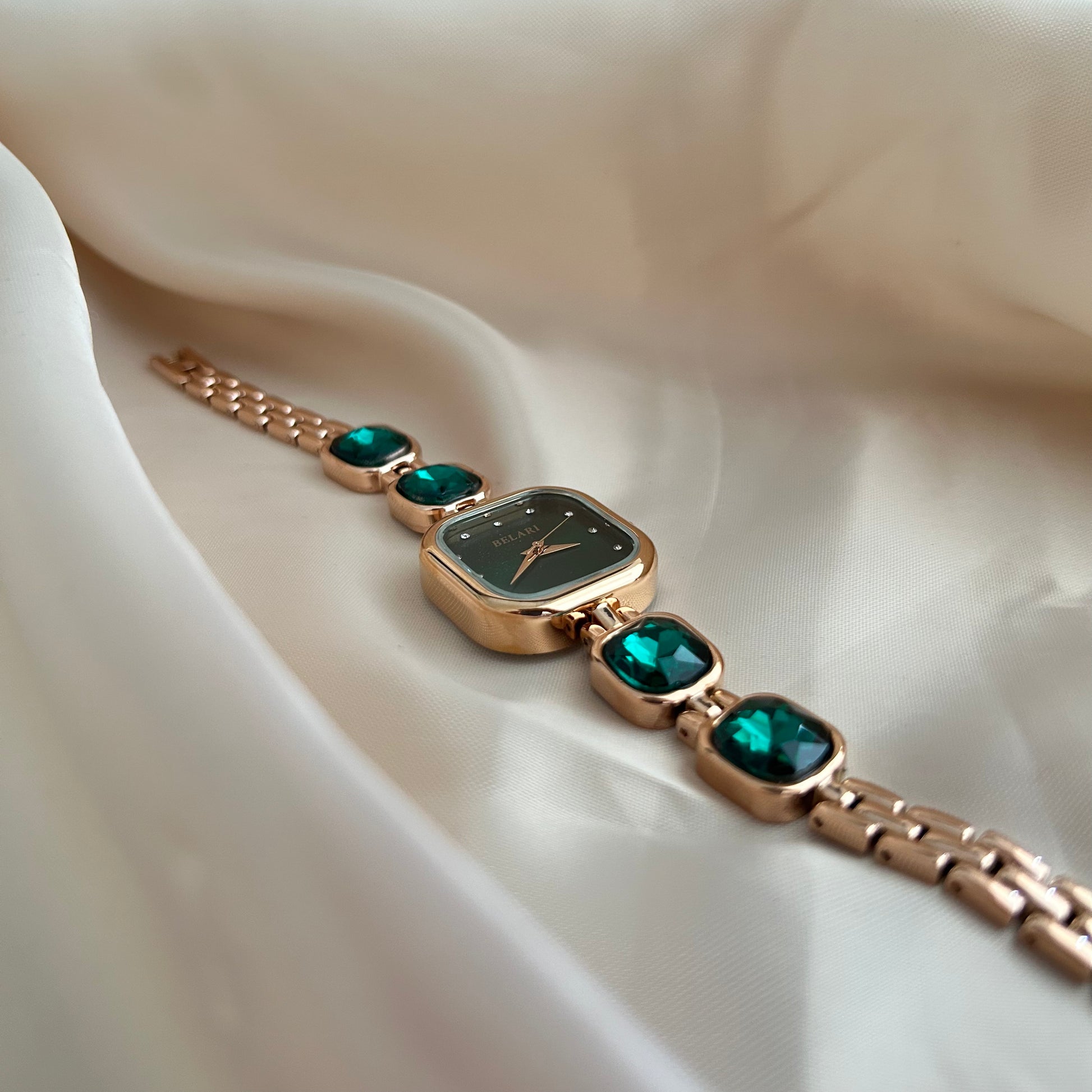 Gold wristwatch with gemstone bracelet links and square dial on satin background.