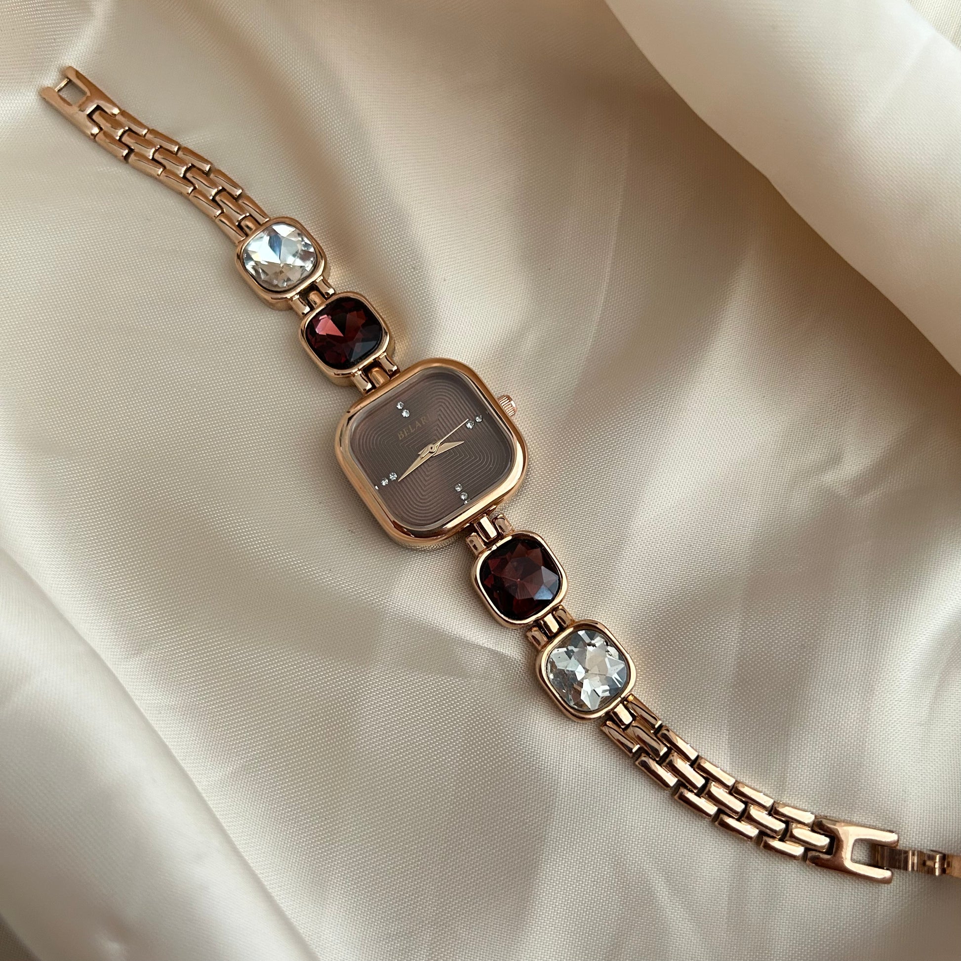 Gold wristwatch with gemstone bracelet links and square dial on satin background.