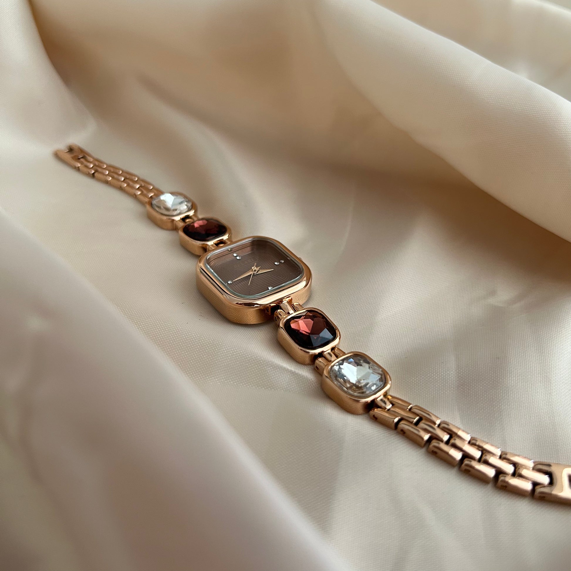 Gold wristwatch with gemstone bracelet links and square dial on satin background.