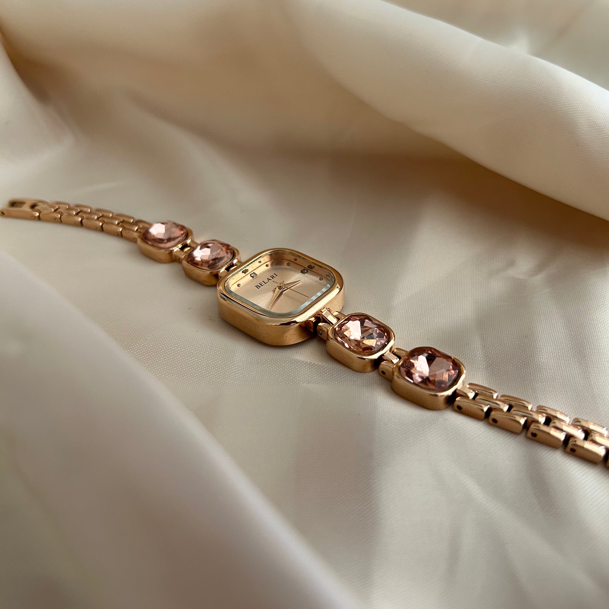 Gold wristwatch with gemstone bracelet links and square dial on satin background.