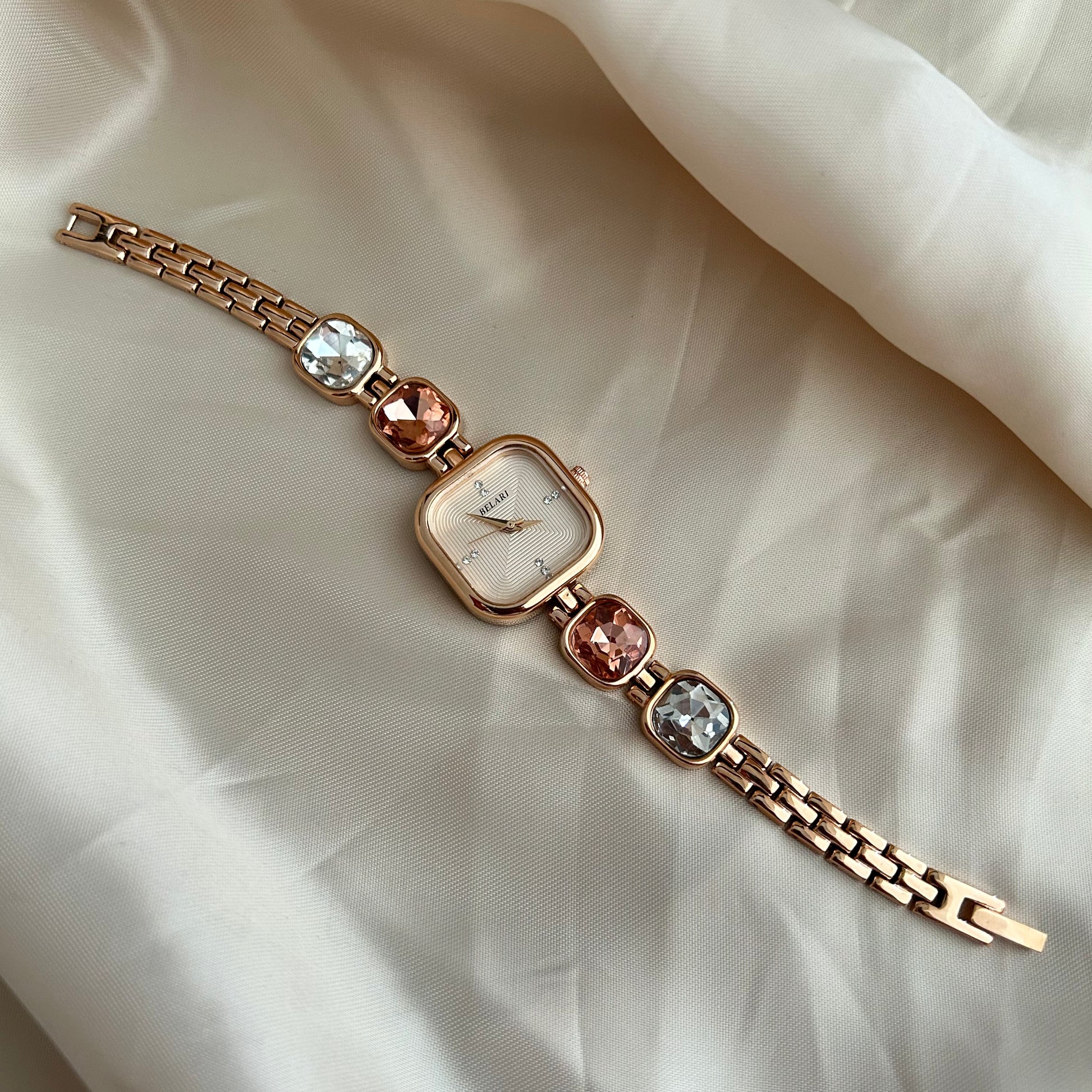 Gold wristwatch with gemstone bracelet links and square dial on satin background.