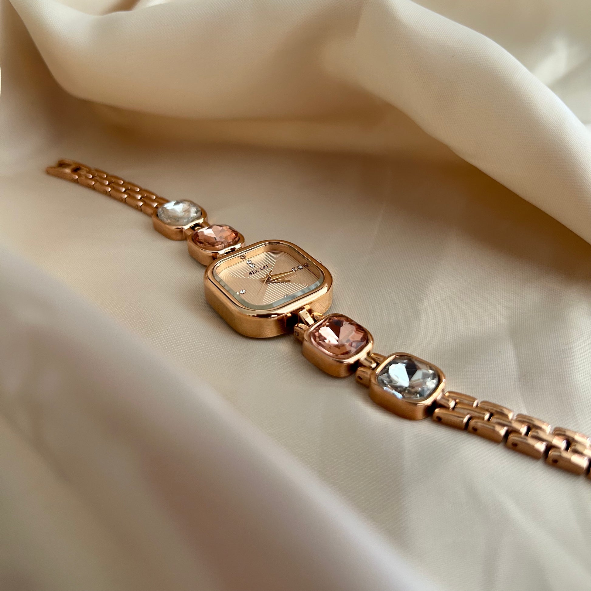 Gold wristwatch with gemstone bracelet links and square dial on satin background.