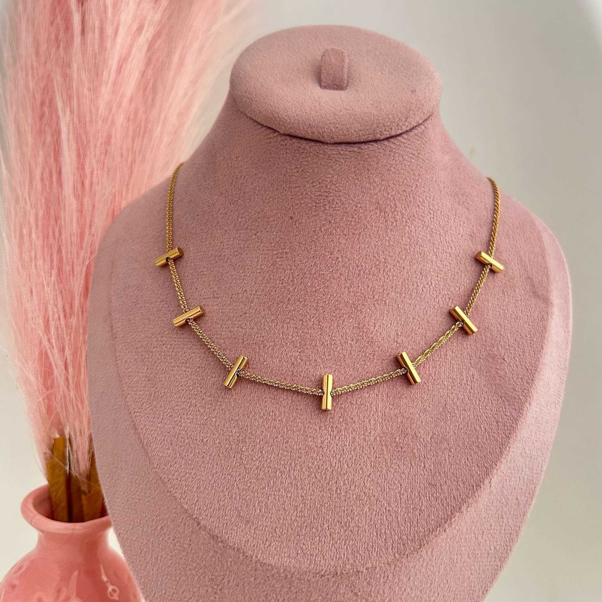 Gold necklace with small horizontal bar charms evenly spaced on chain displayed on pink stand